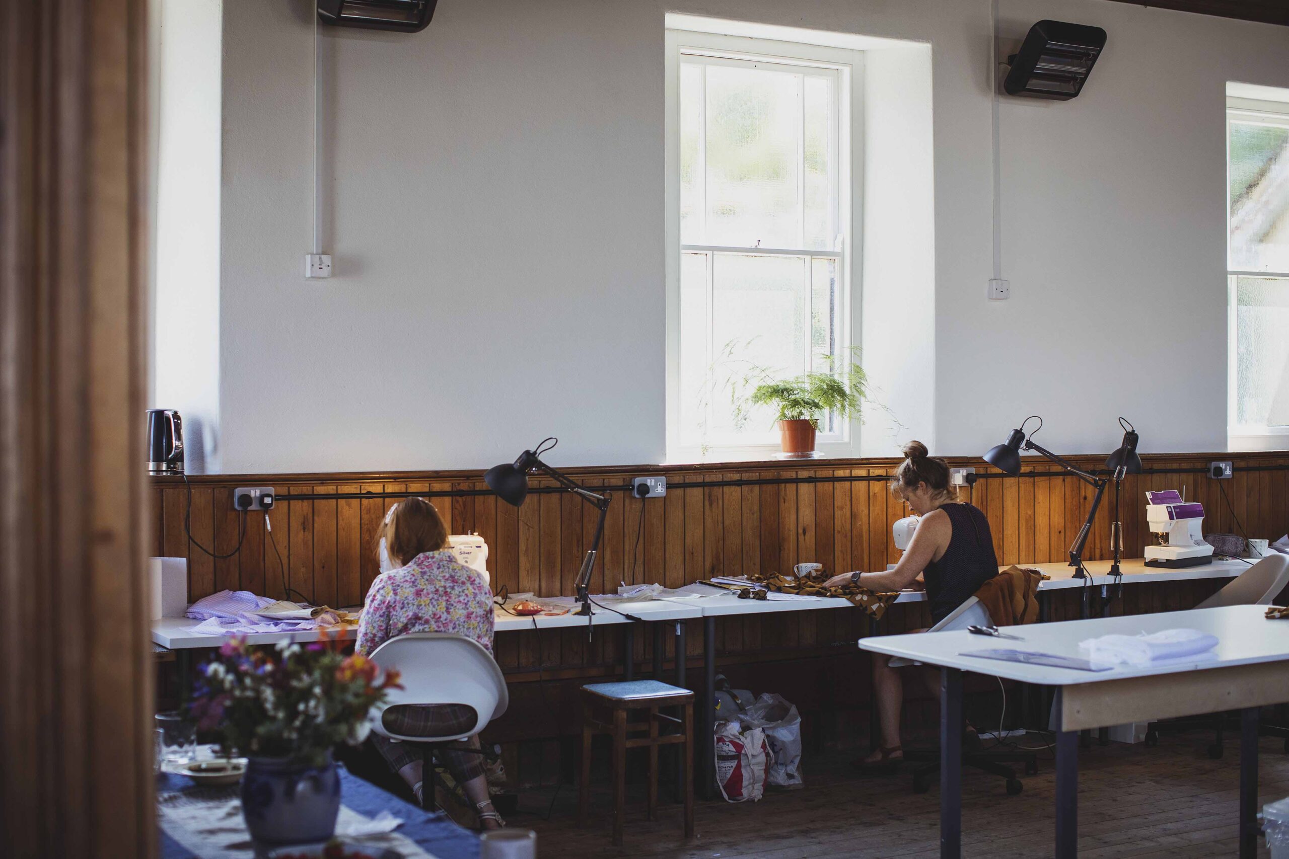 2 sewists sit at their machines in a large sewing studio.